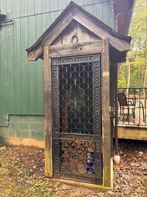 A wooden structure with a decorative metal gate, a small porch with a chair, and a green building in the background.