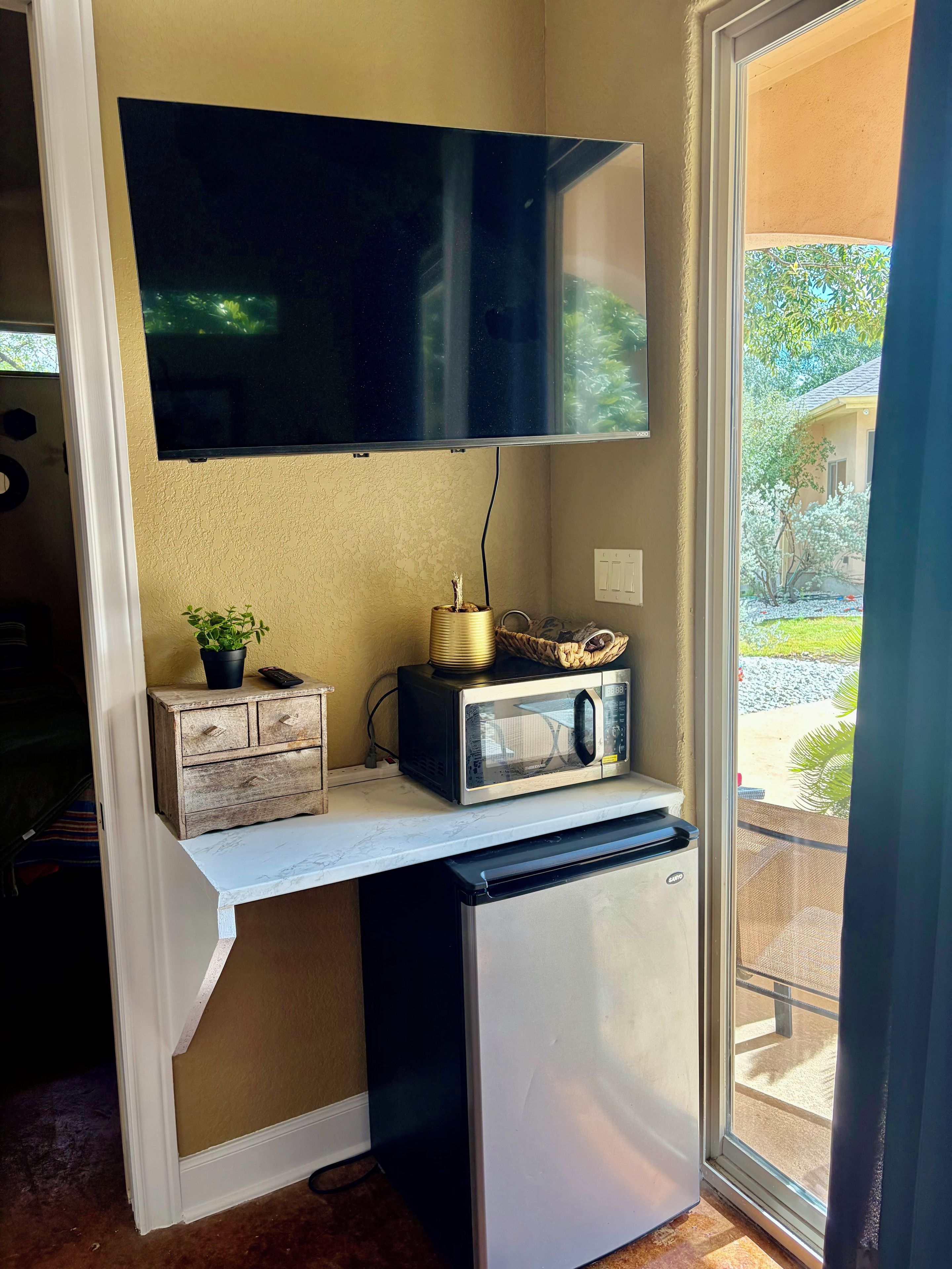 A compact kitchenette with a microwave, a small refrigerator, a television mounted above the counter, and a potted plant on the counter.