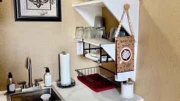 A kitchen with a sink, faucet, and various kitchen items. There is a framed picture on the wall and a stool in front of the sink.