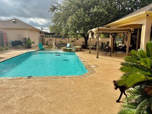 A swimming pool with a lounge area, a covered patio, and a dog in the foreground.