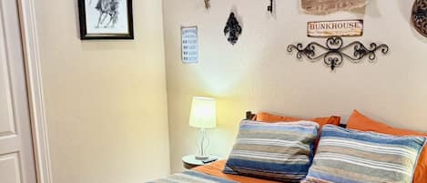 Un dormitorio con cama, almohadas, una mesita de noche, lámpara y adornos en la pared, incluyendo un cartel y un tapiz.