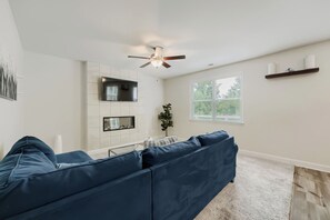 A living room with a blue sofa, a flat-screen TV, a ceiling fan, and a window with blinds. -     Stylish 4BR Home, Fenced Yard, Office + Tesla Charger (Dallas)