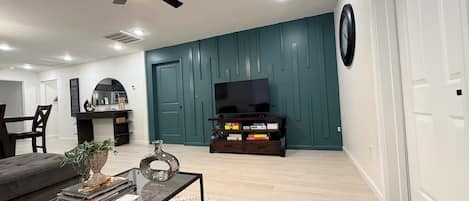 Ein modernes Wohnzimmer mit Deckenventilator, einem Flachbildfernseher auf einem Holzboden, einem Couchtisch mit Büchern und einer Vase sowie einem gemusterten Teppich.