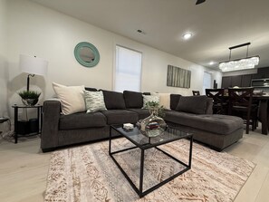 Ein modernes Wohnzimmer mit einem Ecksofa, einem Glastisch und einem Essbereich mit einer Kronleuchter.