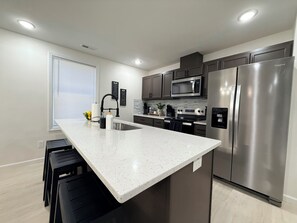 A modern kitchen with a central island, stainless steel appliances, and a black bar stool. - The 5th Street Retreat - Private Fenced Yard (Smithfield)