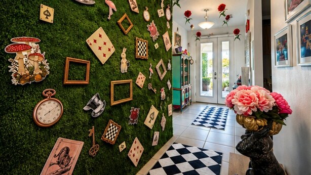Um corredor com parede verde decorada com diversos objetos como relógios, chaves e cartas de baralho. Há rosas vermelhas penduradas no teto e um vaso com flores no final do corredor.