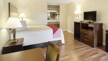 A hotel room with a bed, bedside table, lamp, TV, and a desk with a chair.