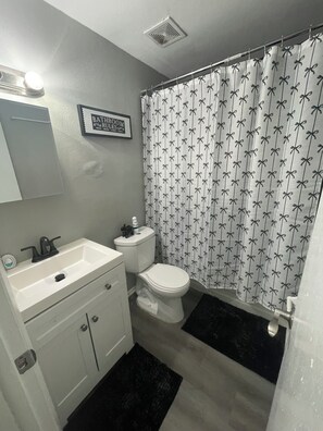 Ein Badezimmer mit einem weißen Waschbecken, einer Toilette, einem Duschvorhang mit Schleifenmuster und einem Schild über dem Waschbecken mit der Aufschrift 'BADEZIMMERREGELN'.