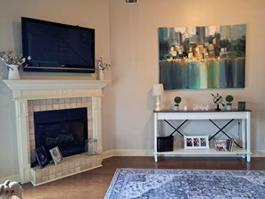 A living room with a fireplace, a television mounted above it, a console table with decorative items, and a large abstract painting on the wall. - Lakefront Auburn Game Day Retreat (Auburn)