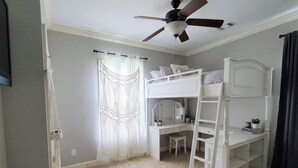 Et værelse med køjeseng, skrivebord, stol, ventilator og et vindue med gardiner.