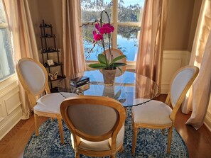 Een eethoek met een ronde glazen tafel, twee houten stoelen met beklede zittingen, een vaas met roze bloemen en een doos kaarten.