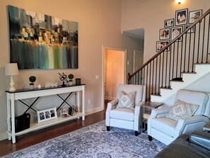 Een woonkamer met een witte console tafel, twee fauteuils, een kamerbreed tapijt, een trap en een groot abstract schilderij.