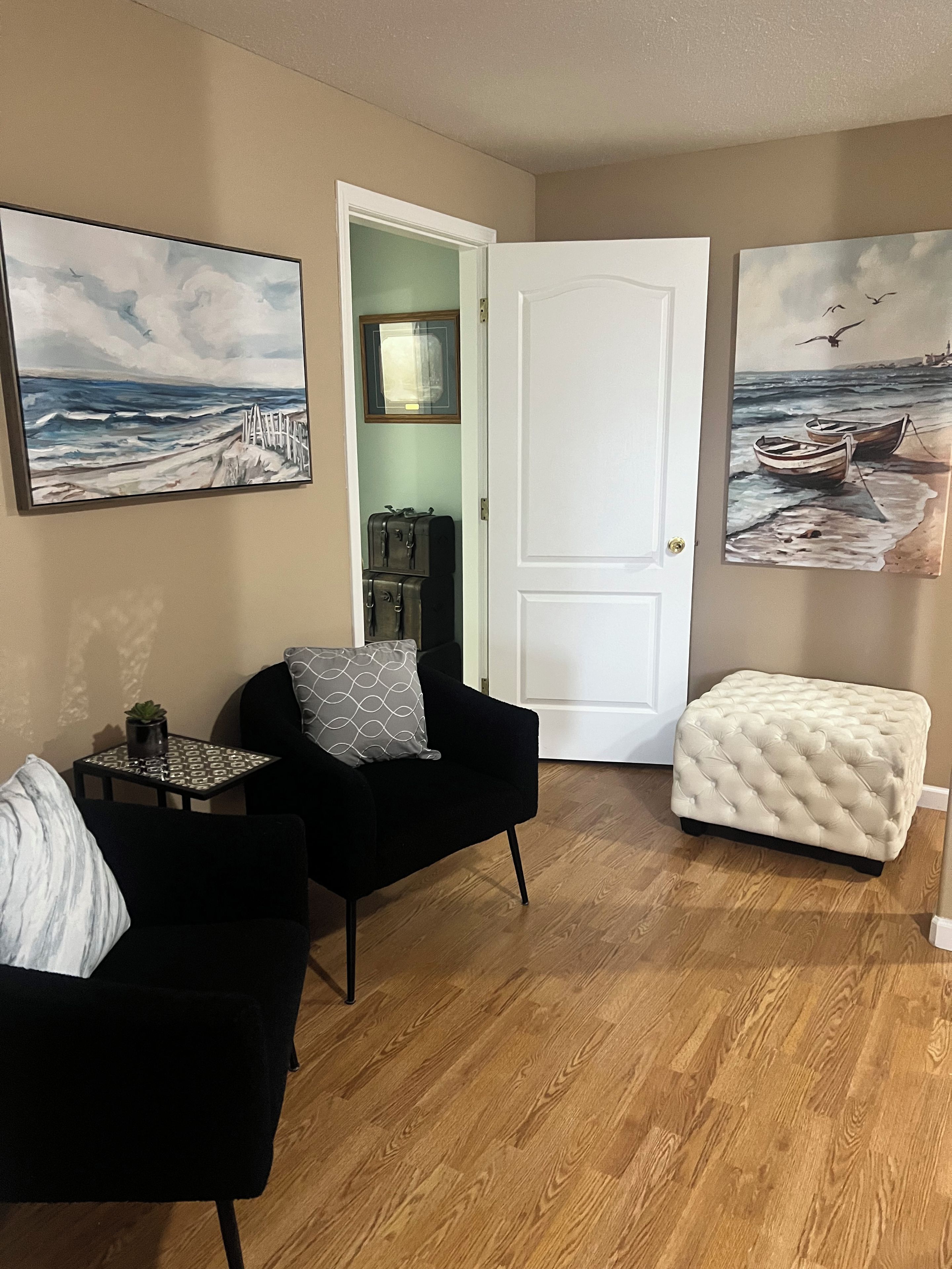 A room with a black armchair, a small table with a plant, a white ottoman, and two paintings depicting seascapes.