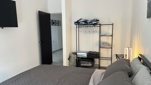 Un dormitorio con una cama, un armario, una estantería con ropa doblada y un televisor en la pared.