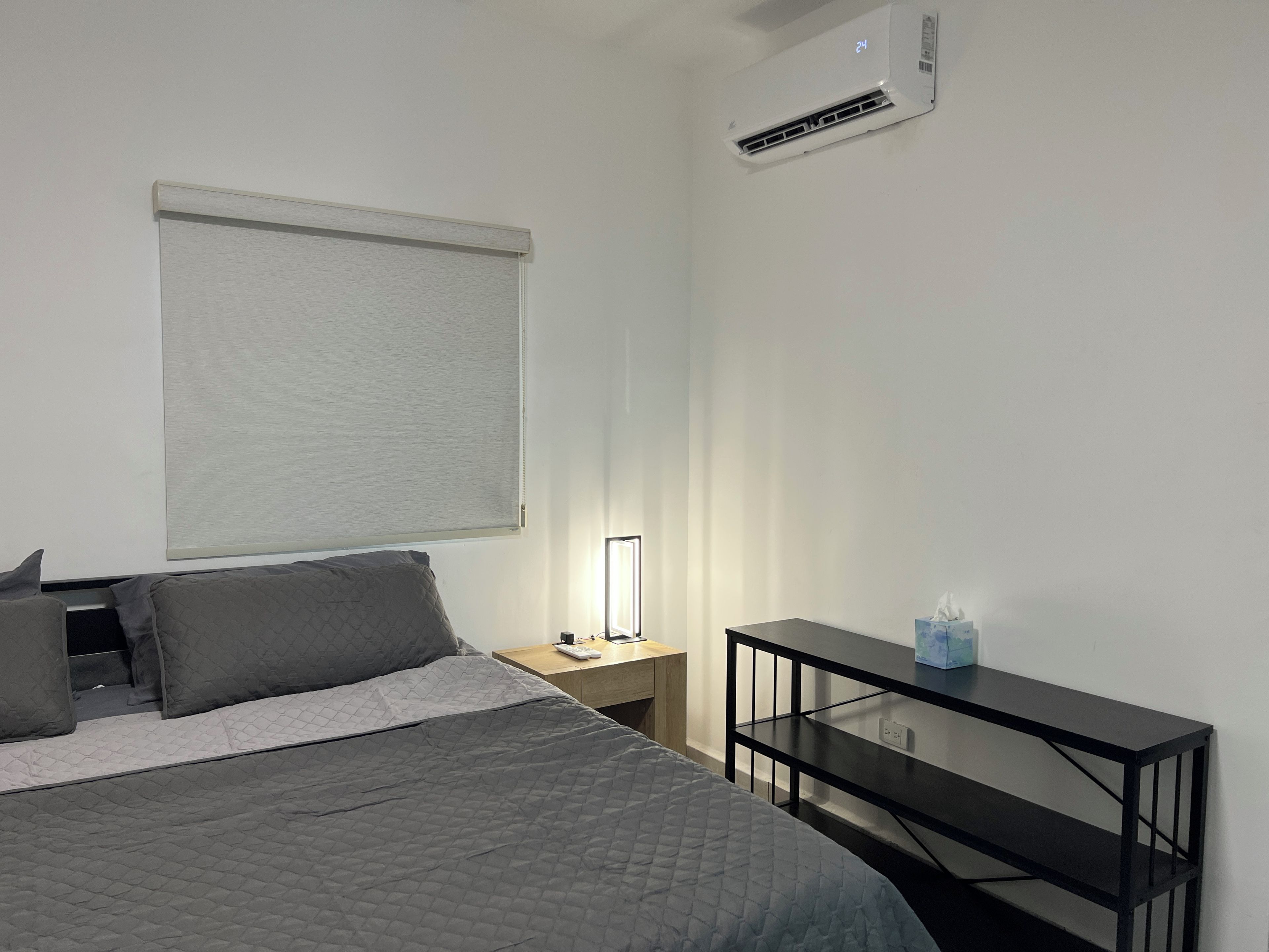 A bedroom with a bed, bedside table, lamp, and air conditioning unit.