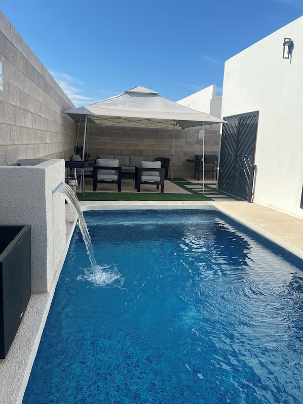 A swimming pool with a water feature, outdoor seating, and a covered area.