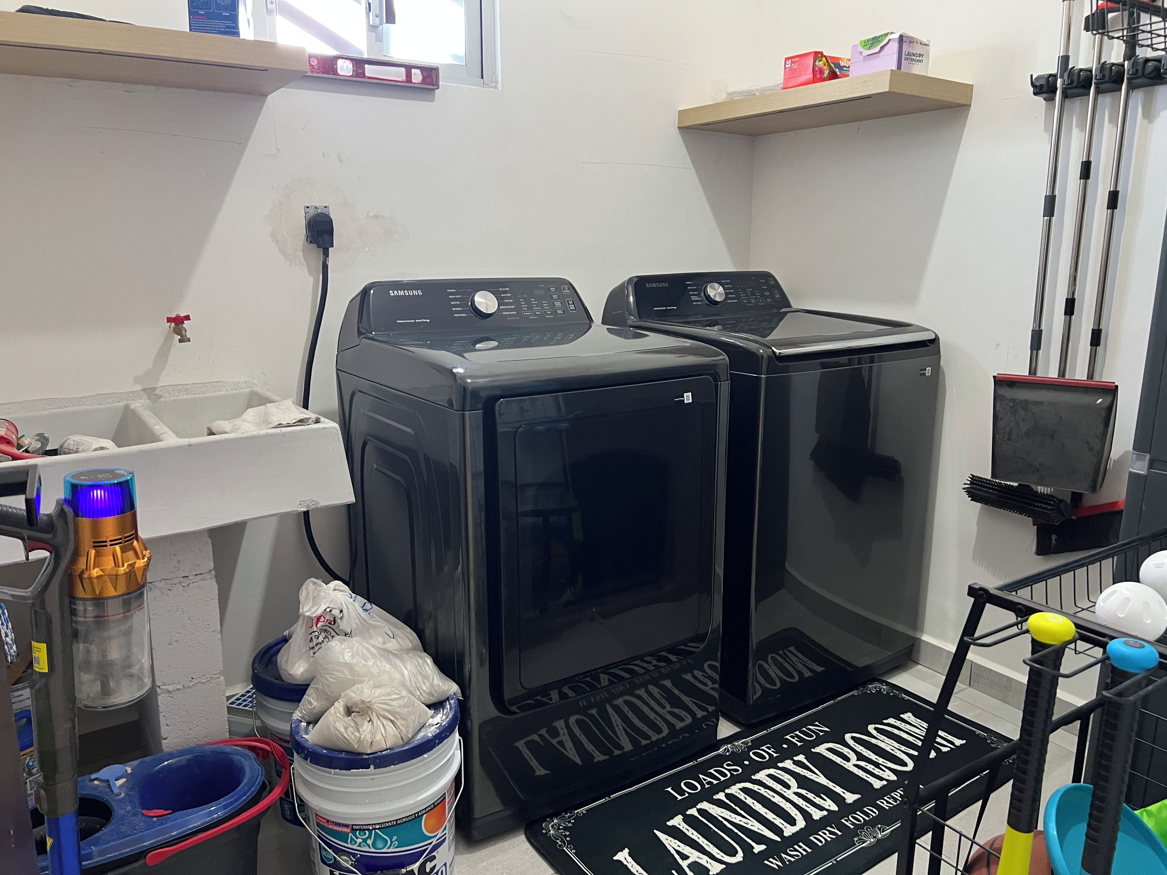 Een wasserette met twee zwarte wasmachines, een mand vol wasgoed en een plank met schoonmaakspullen.