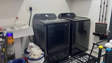 Een wasserette met twee zwarte wasmachines, een mand vol wasgoed en een plank met schoonmaakspullen.