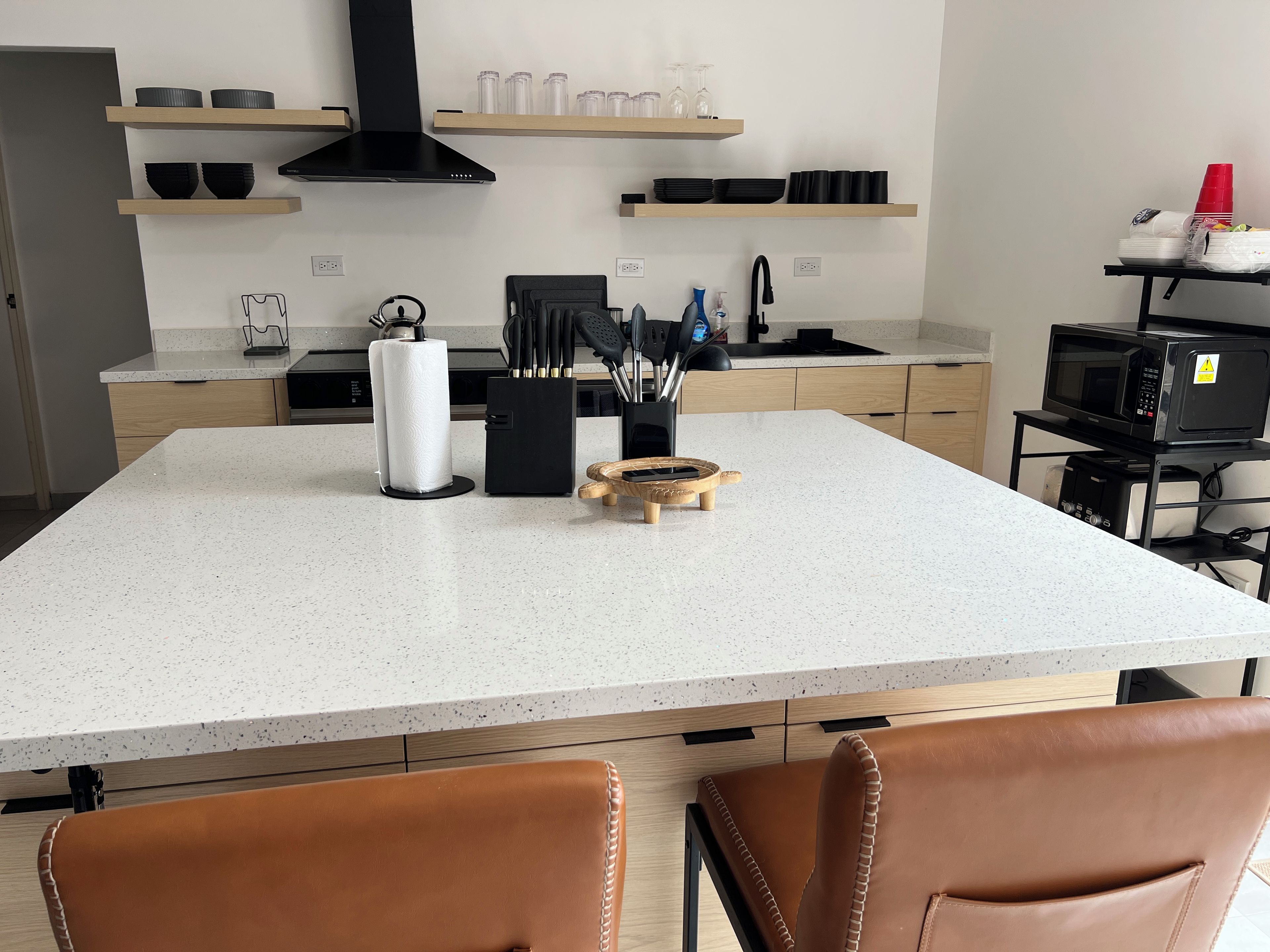 A modern kitchen with a large island, wooden cabinets, and a black microwave on a cart.