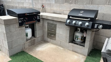 Et udendørs grillområde med to gasgrill, en gasbeholder og et metalskab med skuffer.