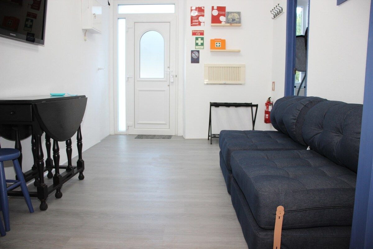 Una habitación con un sofá oscuro, una pequeña mesa con una silla, una puerta blanca y señales de salida de emergencia en la pared.