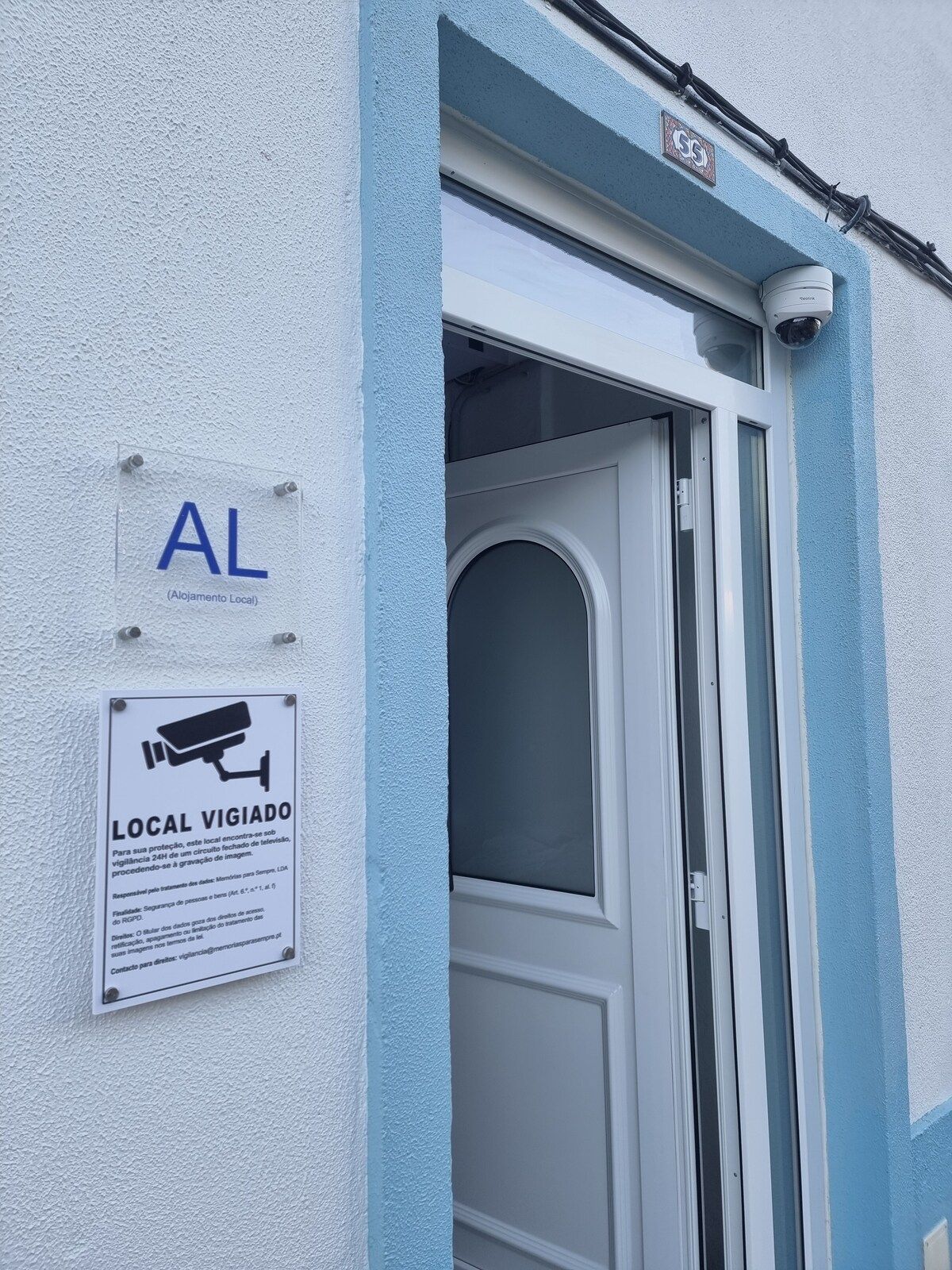 Entrada d'un edifici amb una porta blava, un senyal de càmera de seguretat i un indicador de sistema de vigilància local.