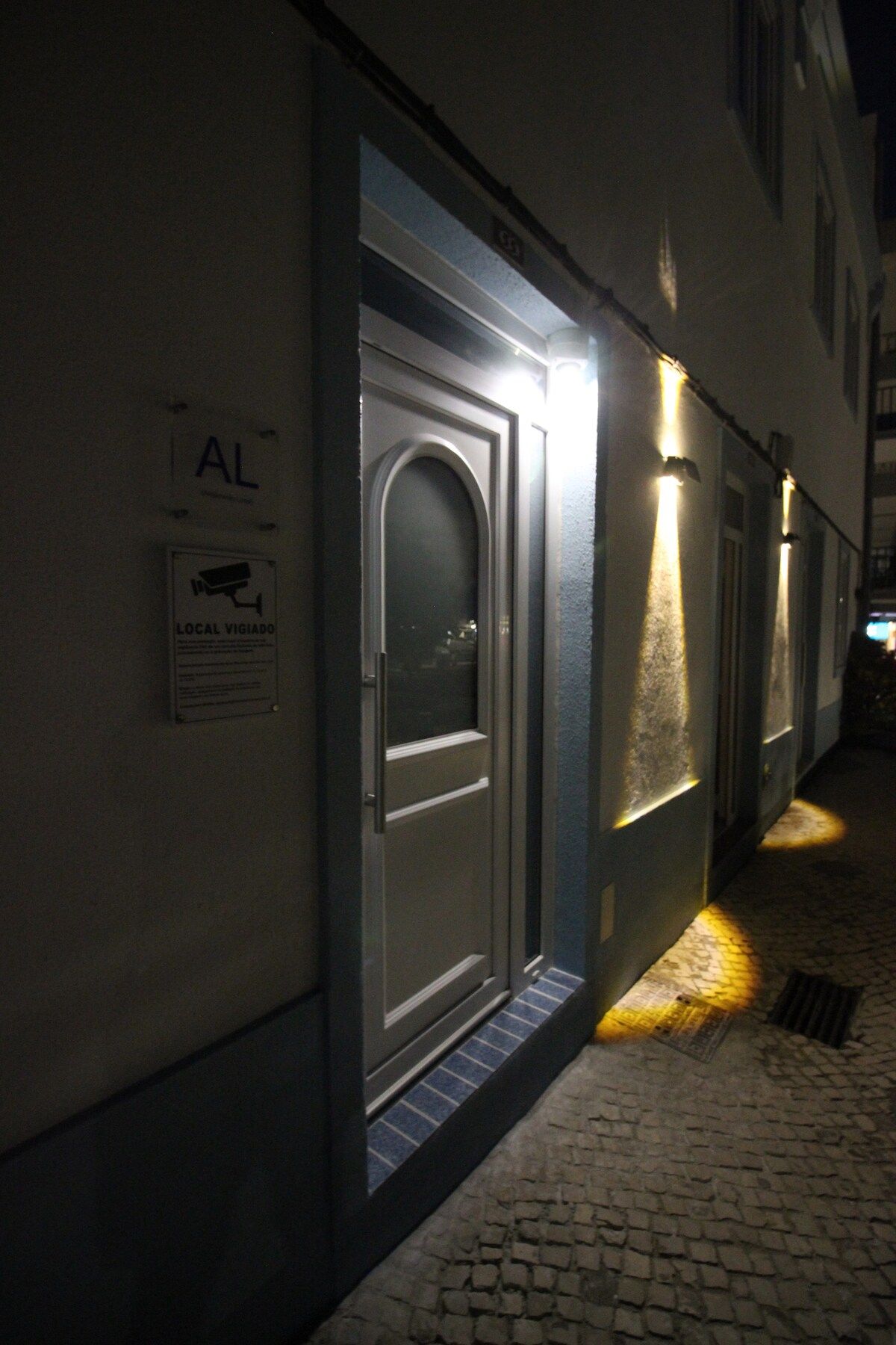 Un callejón estrecho con una puerta iluminada por una tira de luz. Un letrero encima de la puerta dice 'AL' y otro debajo indica 'LOCAL VIGIADO'.