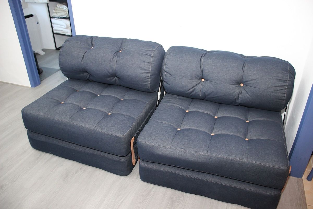 Ein Zimmer mit einer grauen Sofagarnitur, einer weißen Wand, einem Holzboden und einer Tür, die in ein weiteres Zimmer führt.