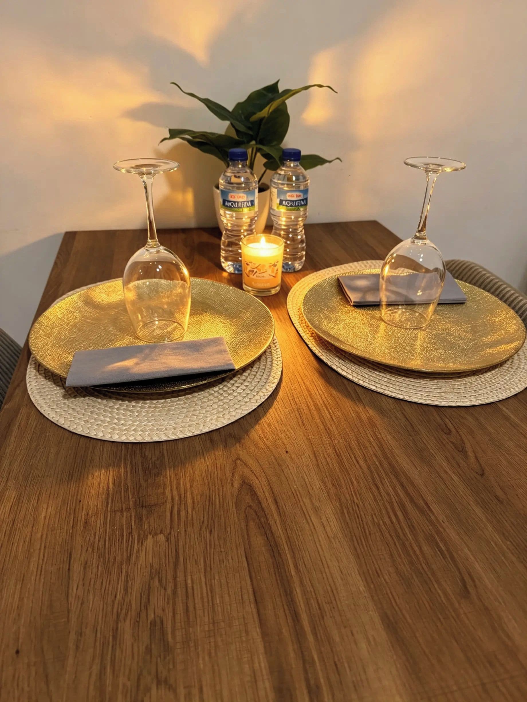 Una mesa de comedor con dos posavasos redondos con detalle dorado, una vela y dos copas de cristal transparentes para vino.