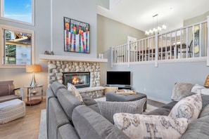 A modern living room with a stone fireplace, a large sofa, a flat-screen TV, and a staircase with a railing. - Ironwood 16 - 4 Bedroom Ski In Ski Out Next to the Gondolas + EZCancel (Steamboat Springs)
