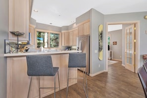 Une cuisine dotée d’un îlot central, de deux tabourets de bar, de meubles en bois, d’un réfrigérateur en acier inoxydable et d’une fenêtre donnant sur la verdure.
