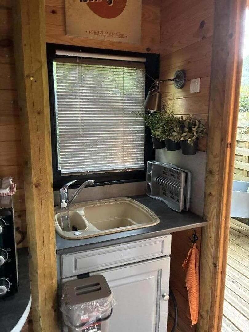 Une cuisine de taille réduite, équipée d’un évier, d’une fenêtre avec stores, d’un radiateur et d’un petit placard.