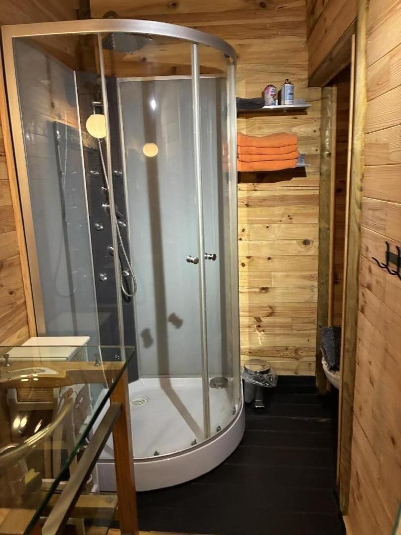 Une salle de bain rustique, avec une cabine en bois, une douche en verre, un lavabo et une petite étagère contenant des articles de toilette.