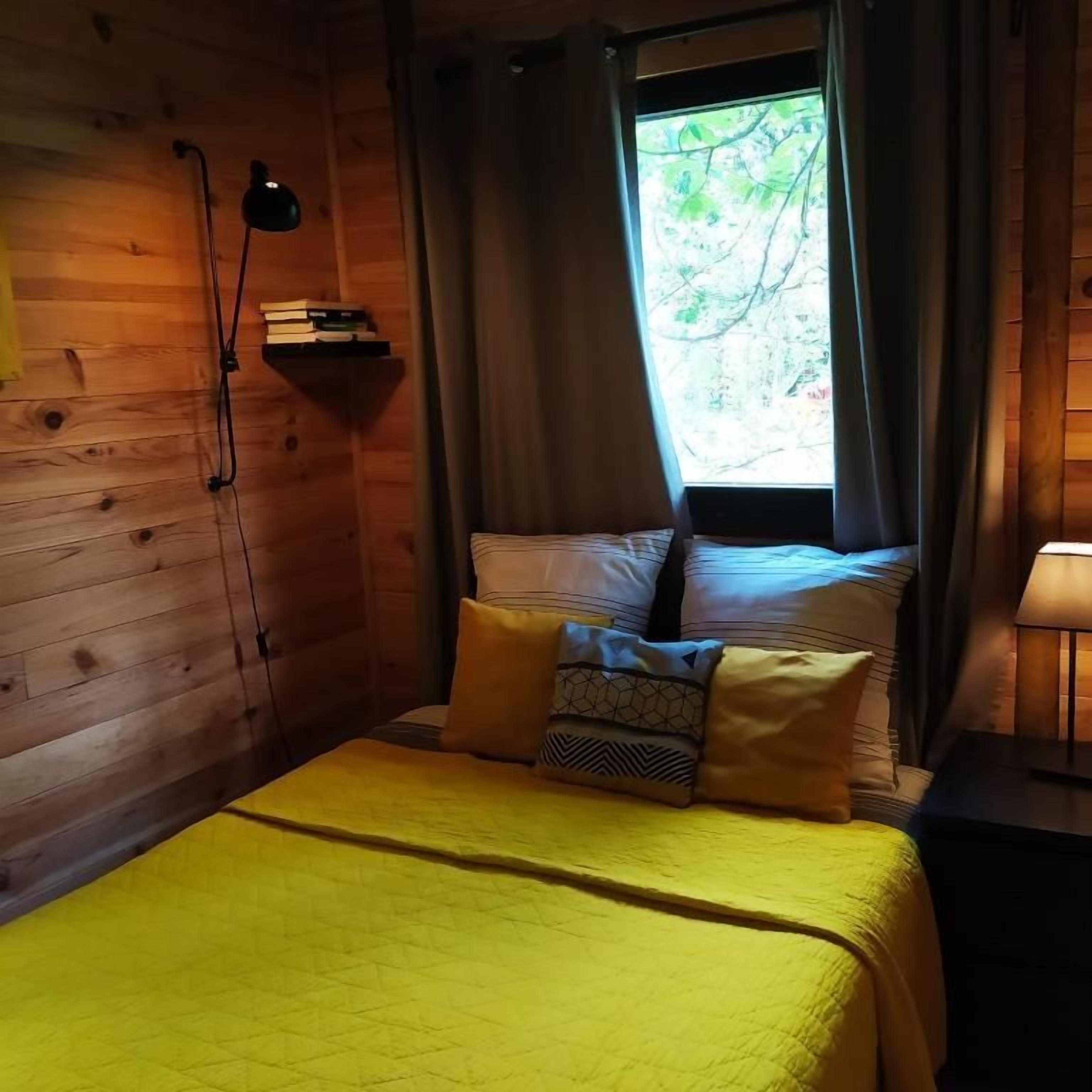 Une pièce de style cabane en bois, comprenant un lit, une table de chevet, une lampe, une étagère avec des livres et une fenêtre avec des rideaux.