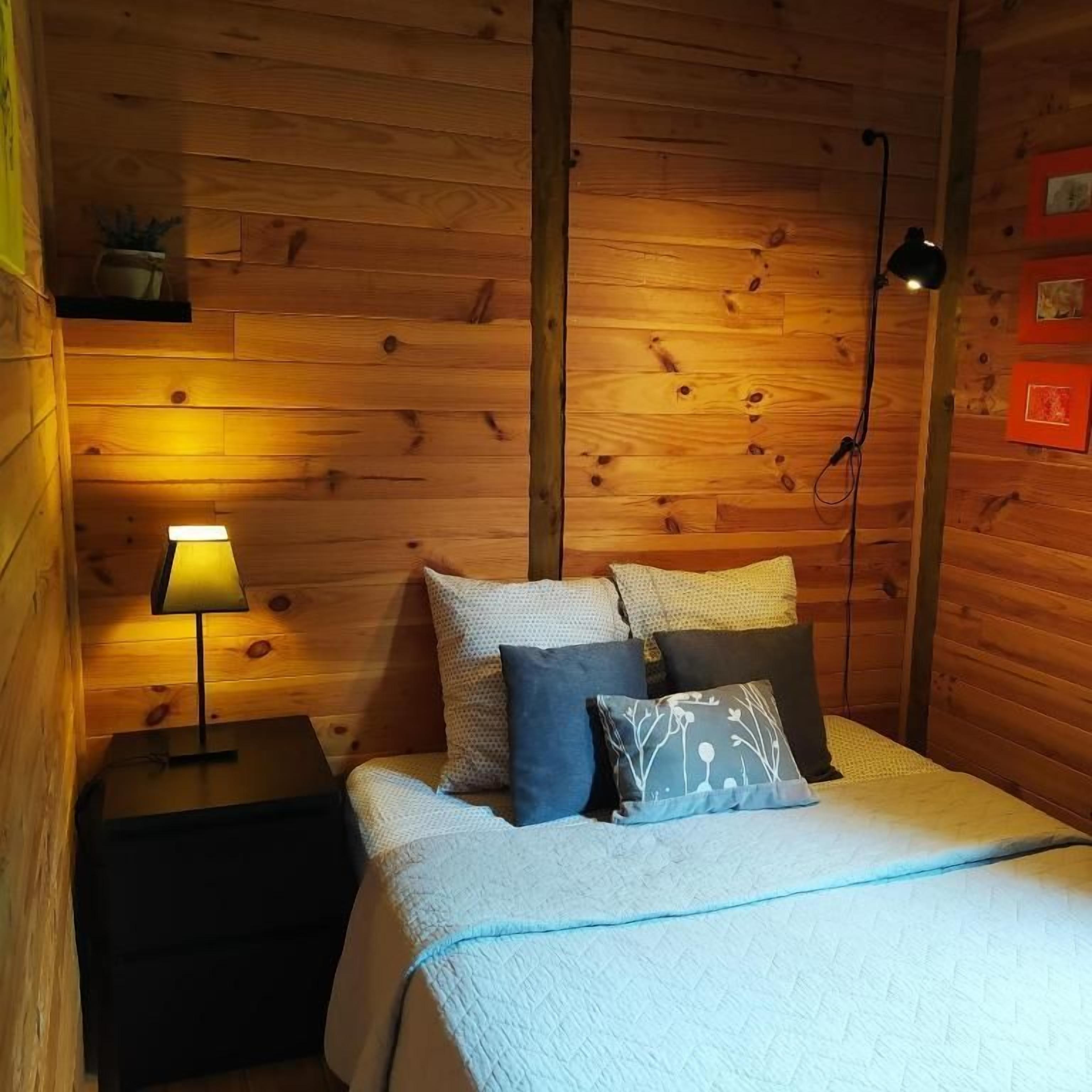 Une pièce aux murs lambrés, comprenant un lit, une table de chevet, une lampe et une étagère fixée au mur, sur laquelle se trouve une plante.