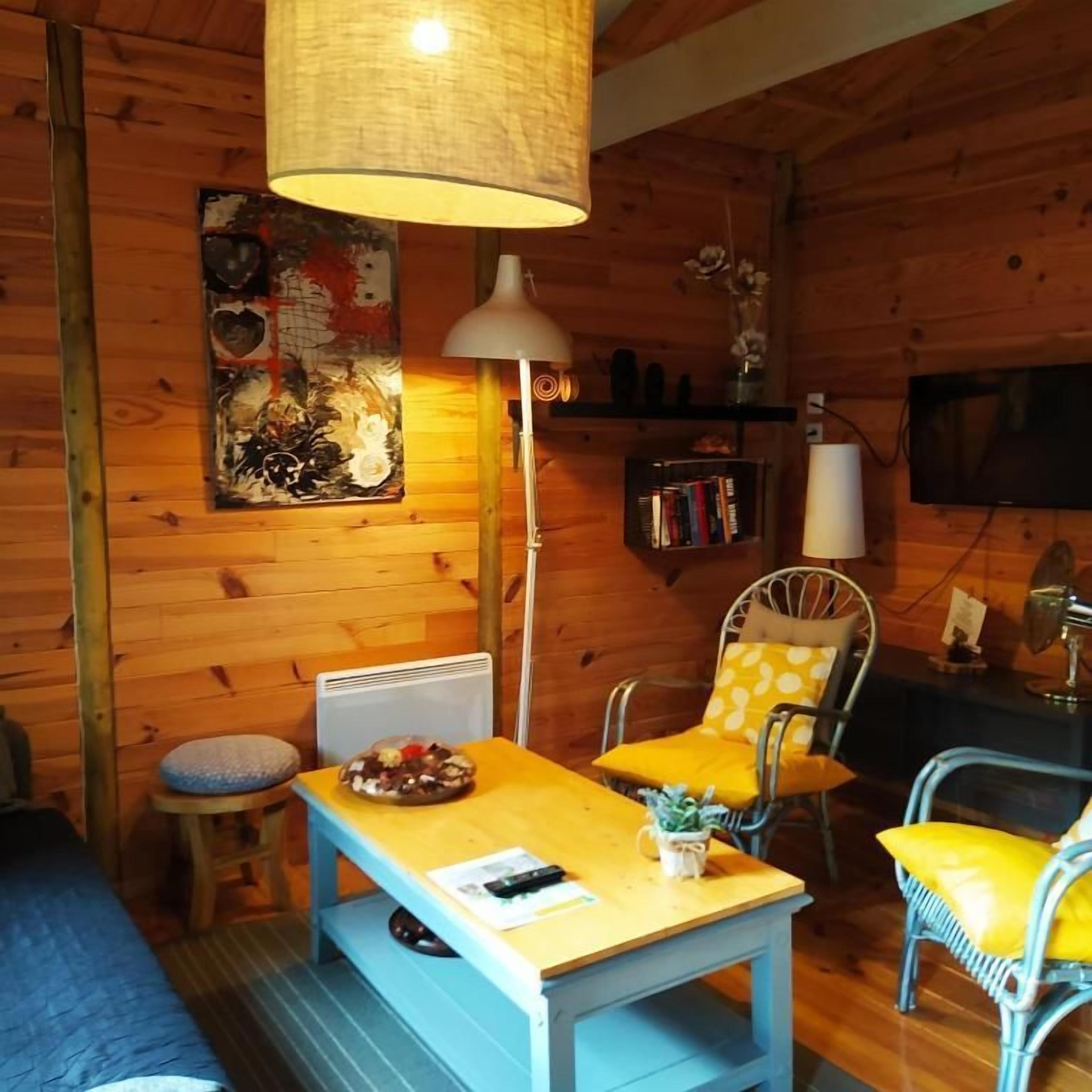 A cozy cabin interior with a wooden wall, a dining table, two chairs, a sofa, a lamp, a bookshelf, and a television.