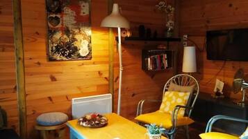 A cozy cabin interior with a wooden wall, a dining table, two chairs, a sofa, a lamp, a bookshelf, and a television.