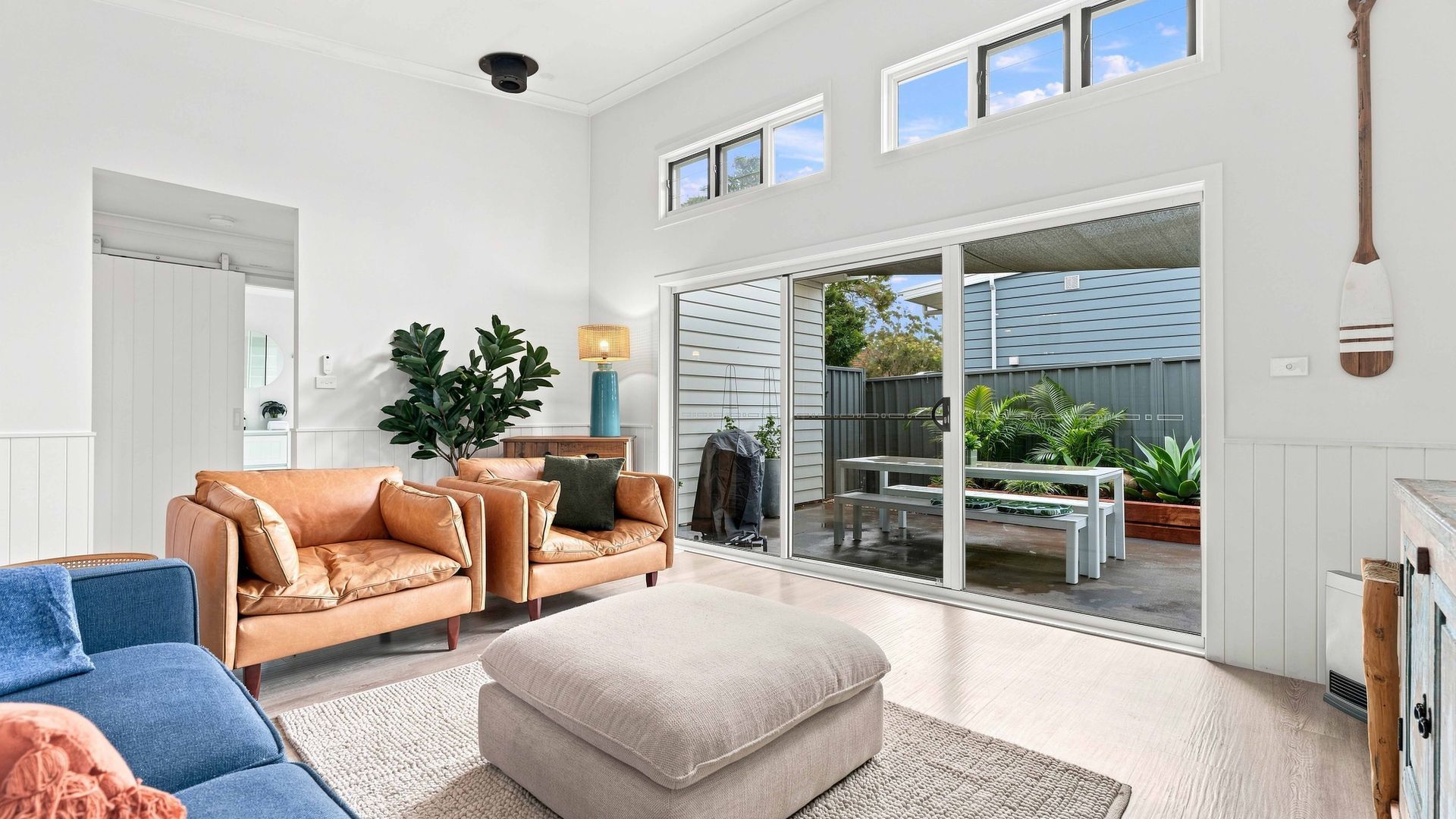 A spacious living room with a leather sofa, a blue armchair, and a neutral-toned ottoman. A large sliding glass door leads to an outdoor patio area with a table and chairs.