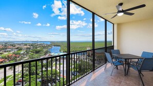 A balcony with a table and chairs, a ceiling fan, and a view of a coastal area with buildings and greenery.