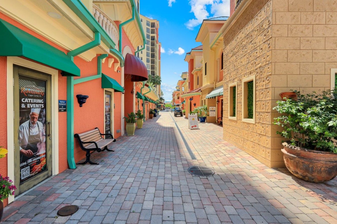 A paved walkway flanked by buildings with colorful facades, a bench, and potted plants.