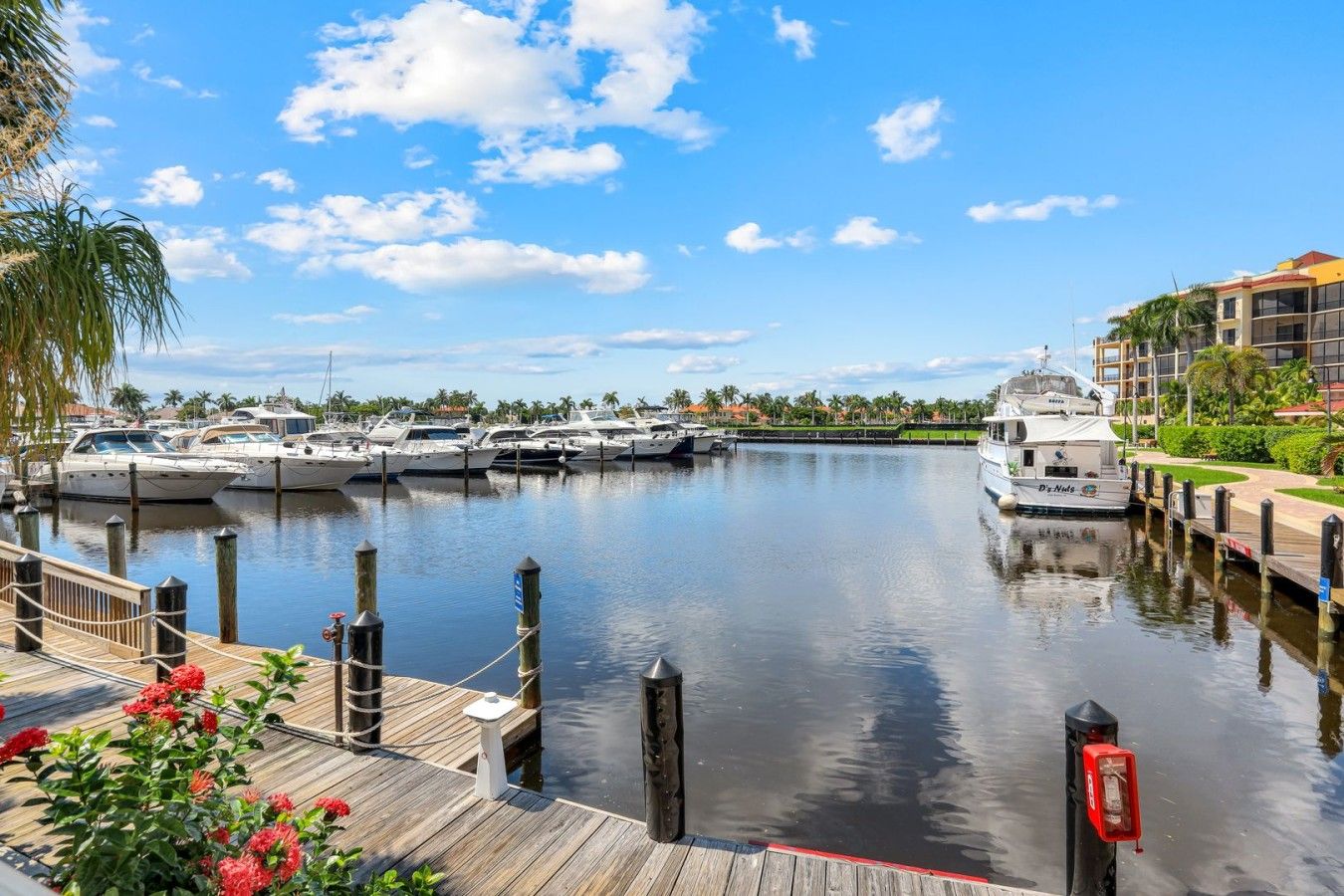 A marina with several yachts docked, a wooden dock with red flowers, and a building with a yellow roof in the background.