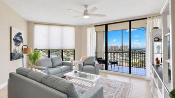 A spacious living room with a balcony, a ceiling fan, and a large window offering a city view.