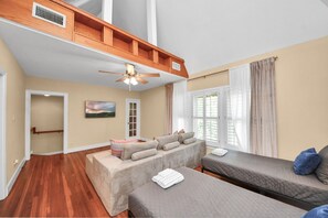 Una habitación con cama alta, un sofá y una cama. Tiene ventilador de techo, una ventana con cortinas y una puerta.