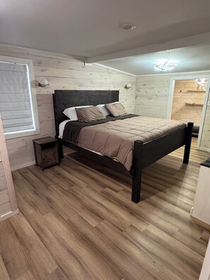 Une chambre avec un plancher en bois, un lit doté d’une tête de lit sombre, une table de chevet et une fenêtre avec des stores.