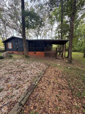 Une cabane dotée d’une terrasse en bois et d’un porche couvert, entourée d’arbres et bordée d’une zone recouverte de gravier.