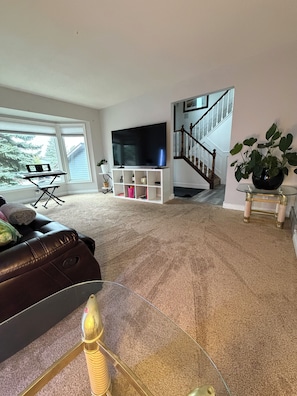 A living room with a large flat-screen TV, a leather sofa, a glass coffee table, and a staircase in the background. - Bright & Spotless 3BR plus Free parking Perfect for Families & Long Stays (Calgary)