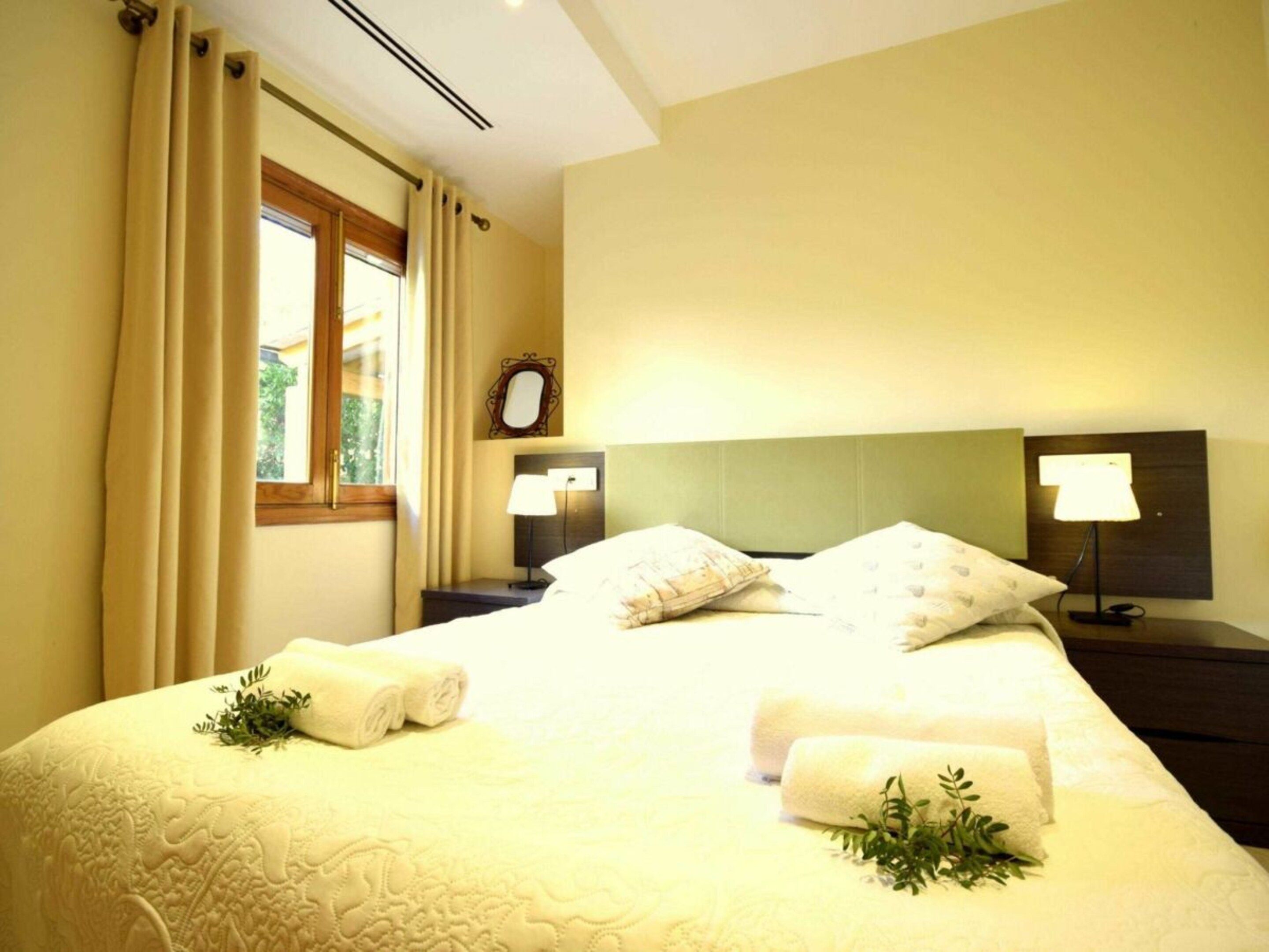 A hotel room with a bed, two bedside lamps, a nightstand, a window with curtains, and a mirror on the wall.