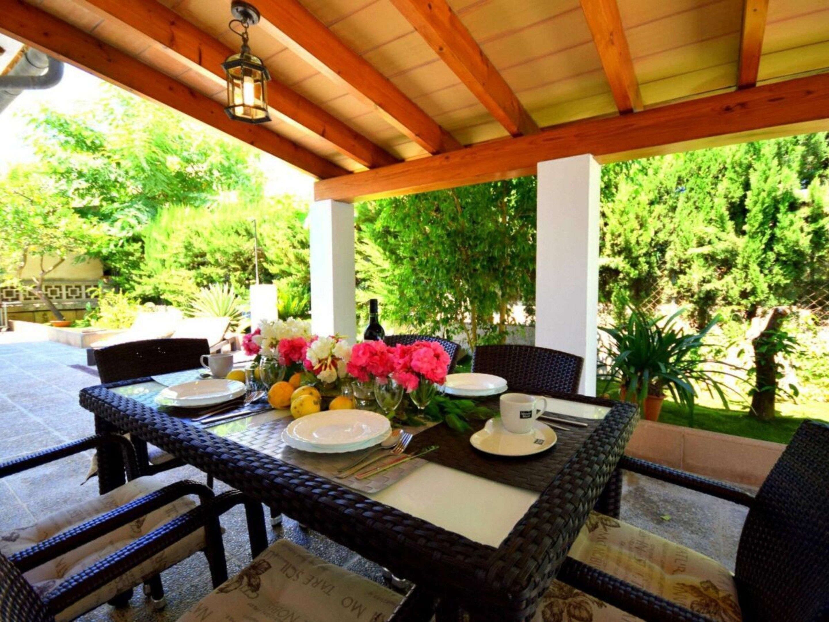 An outdoor patio with a table set for dining, surrounded by wicker chairs and a wooden pergola.