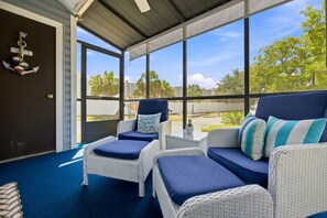A covered outdoor seating area with wicker furniture and cushions, large windows offering views of palm trees and buildings. - Mariner Lakes 14 Serenity & Sandy Shores (Orange Beach)