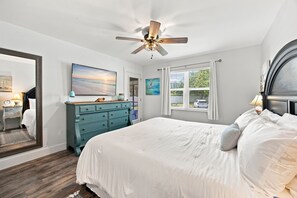 Ein Schlafzimmer mit einem Bett, einem Schminktisch, einem Deckenventilator, einem Bild und einem Fenster mit Vorhängen.
